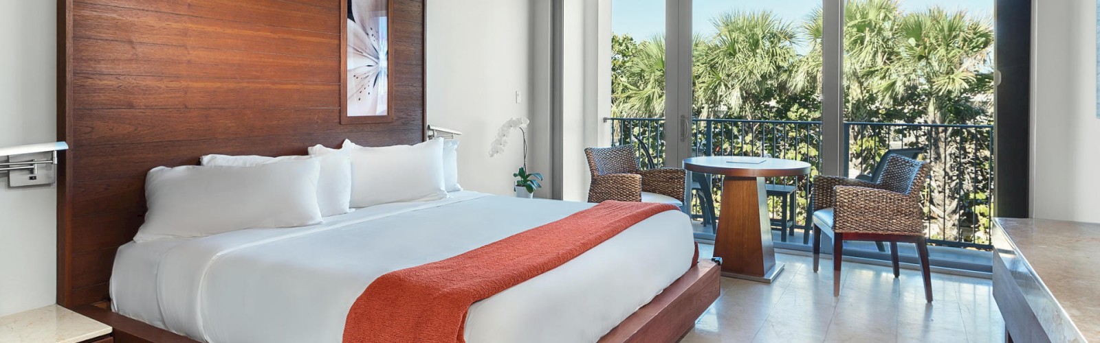 This image shows a modern hotel room with a large bed, an orange throw blanket, a small table with two chairs, and a balcony with a scenic view.