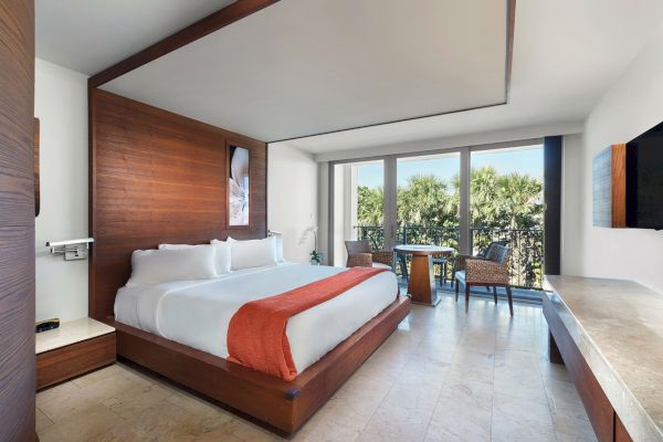 A modern hotel room features a large bed with white linens and an orange throw, a TV, a small table with chairs, and floor-to-ceiling windows.