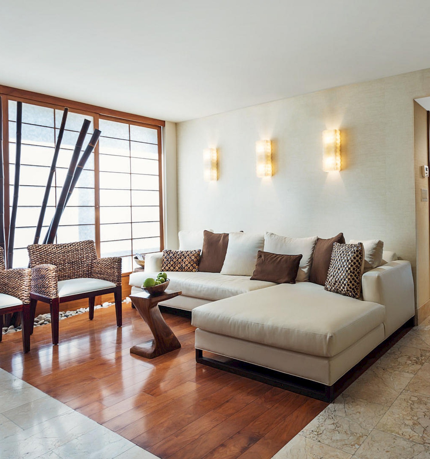 A modern living room features a white sectional sofa, a wooden coffee table, woven chairs, and ambient wall lights, leading to a hallway.