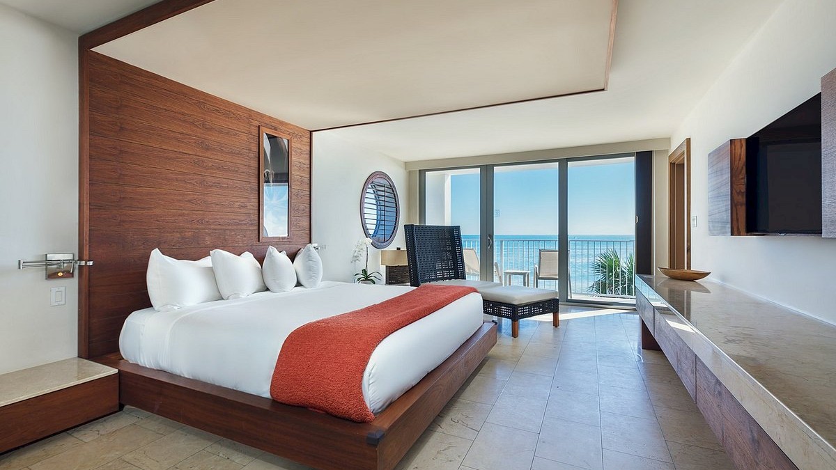 A modern hotel room with a large bed, orange throw, wooden accents, seating area, and a balcony with sea views.