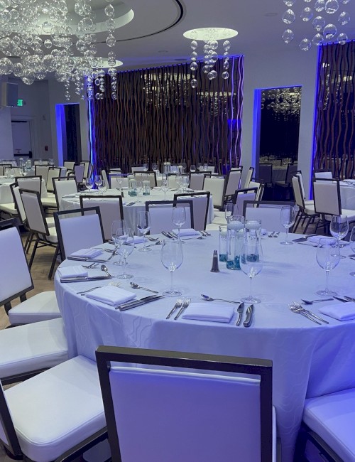 An elegant banquet room with round tables set for dining, adorned with white linens and contemporary lighting fixtures above.