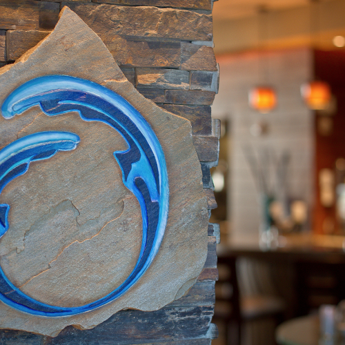 A stone sign with a blue wave design is mounted on a wall. The background shows a warmly lit interior space, possibly a bar or restaurant.
