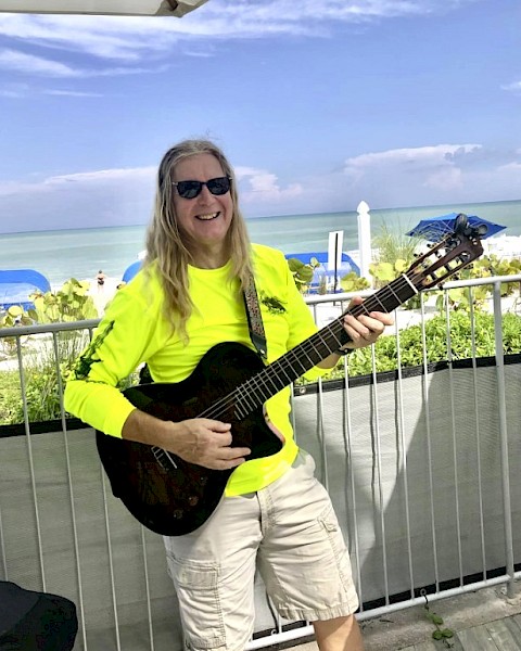 A person in sunglasses and a neon shirt plays guitar by the beach, with blue umbrellas and ocean in the background.