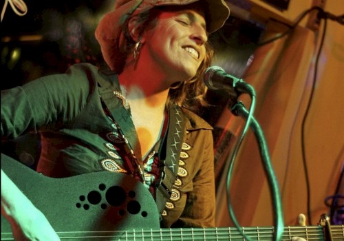 A musician is playing a guitar and singing into a microphone, wearing a hat and smiling during a performance.