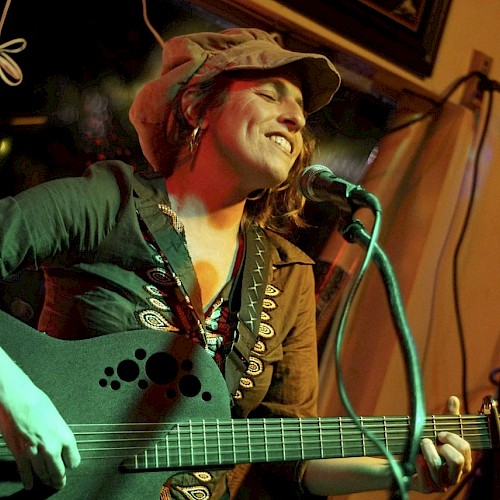 A musician is playing a guitar and singing into a microphone, wearing a hat and smiling during a performance.