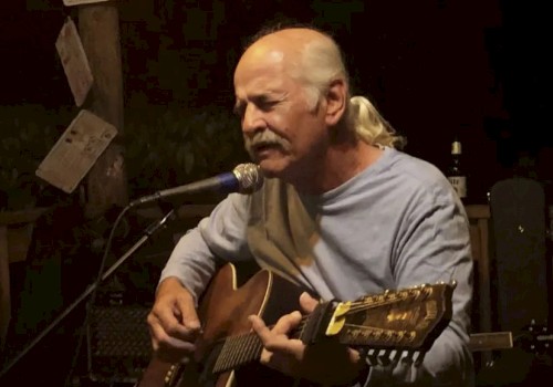 A man with a mustache is playing an acoustic guitar and singing into a microphone, surrounded by a dimly lit, cozy environment.