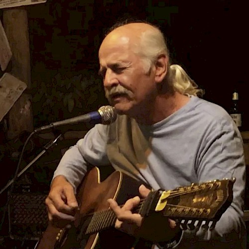 A man with a mustache is playing an acoustic guitar and singing into a microphone, surrounded by a dimly lit, cozy environment.