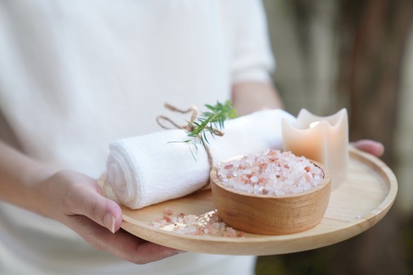 A person holds a wooden tray with a towel, a sprig of rosemary, a bowl of Himalayan salt, and a lit candle for a spa-like experience.