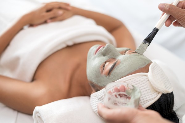 A person is lying down receiving a facial treatment with a green face mask, applied with a brush by another individual.