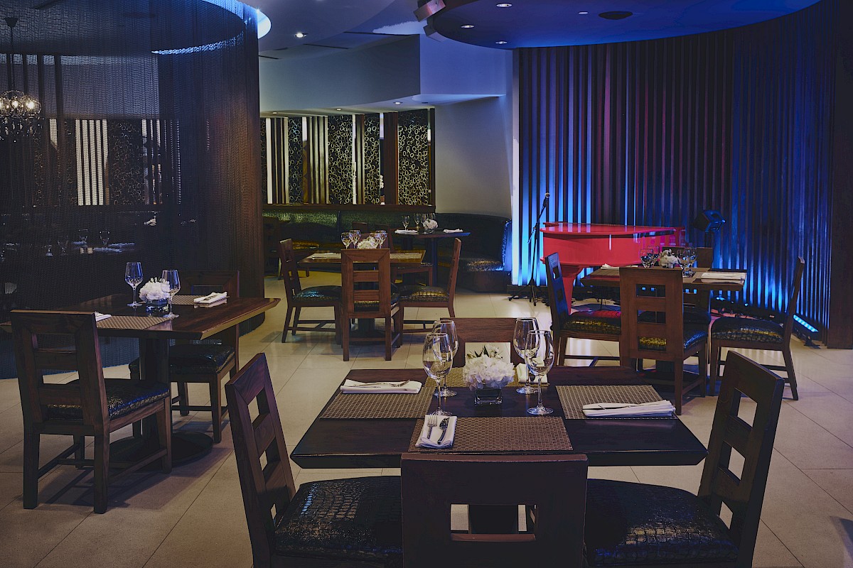 A dimly lit restaurant with wooden tables, chairs, place settings, and ambient blue lighting, creating a cozy atmosphere.