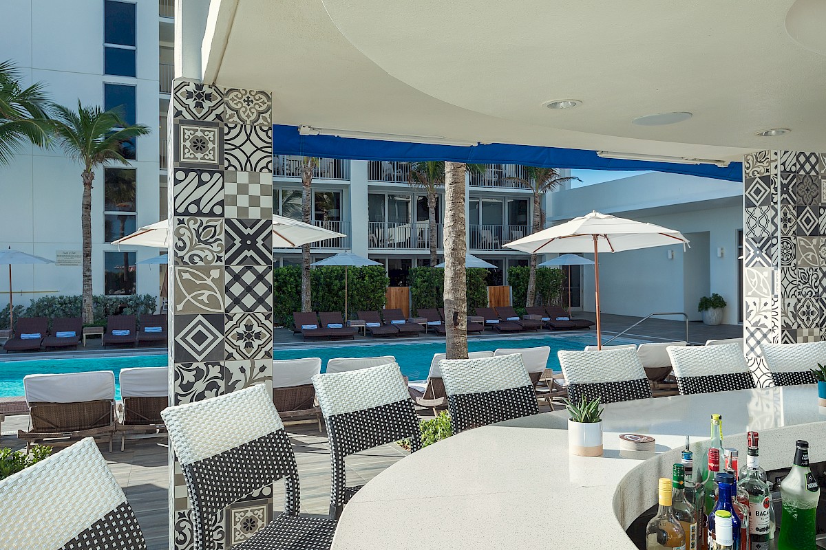 Outdoor bar with patterned pillars, wicker stools, a display of bottles, and a view of a pool with sun loungers and umbrellas.