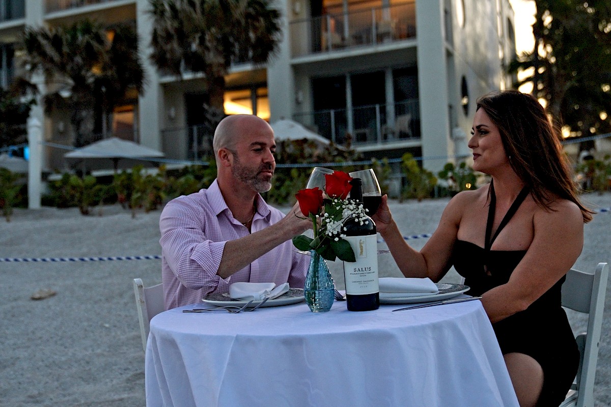A couple is enjoying a romantic dinner at a table set with wine and roses on a beach, in front of a building at sunset.