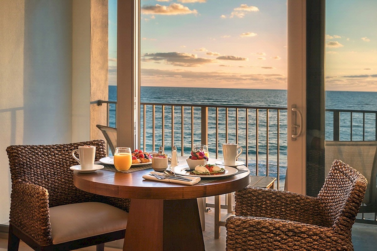 A breakfast setup on a table by a window overlooking the ocean, featuring juice, coffee, fruit, and pastries in a cozy setting.