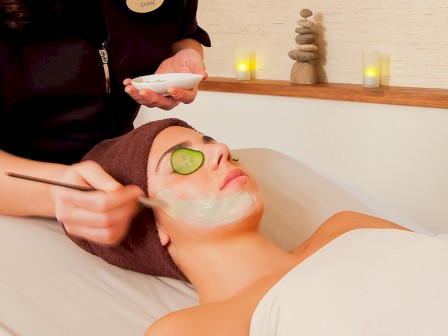 A person receives a facial treatment, lying down with a towel, a face mask, and cucumber on eyes in a spa setting with candles.
