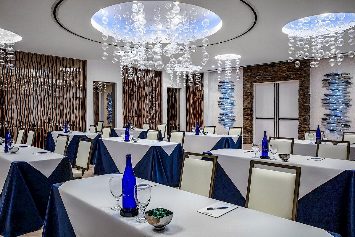 A modern meeting room with tables set with blue bottles, glasses, and notepads, featuring decorative ceilings and wood panel accents.