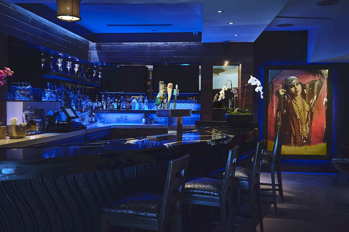 A dimly lit bar with blue lighting, barstools, floral decorations, and a colorful framed artwork in the background.