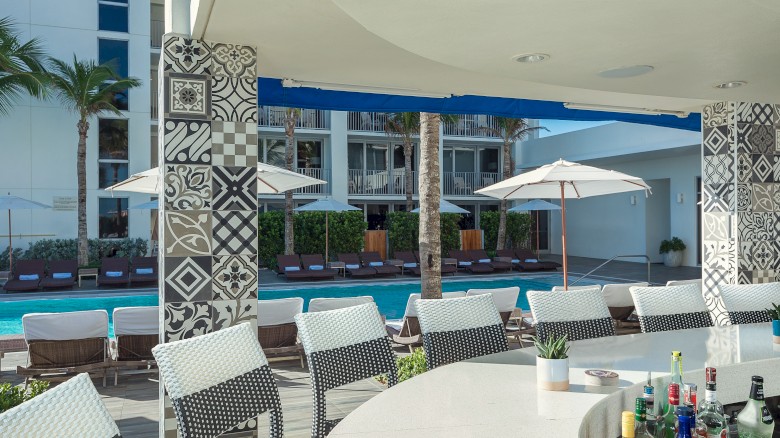 Poolside bar with patterned columns, stools, and liquor bottles. Lounge chairs and umbrellas by the pool, under clear skies.