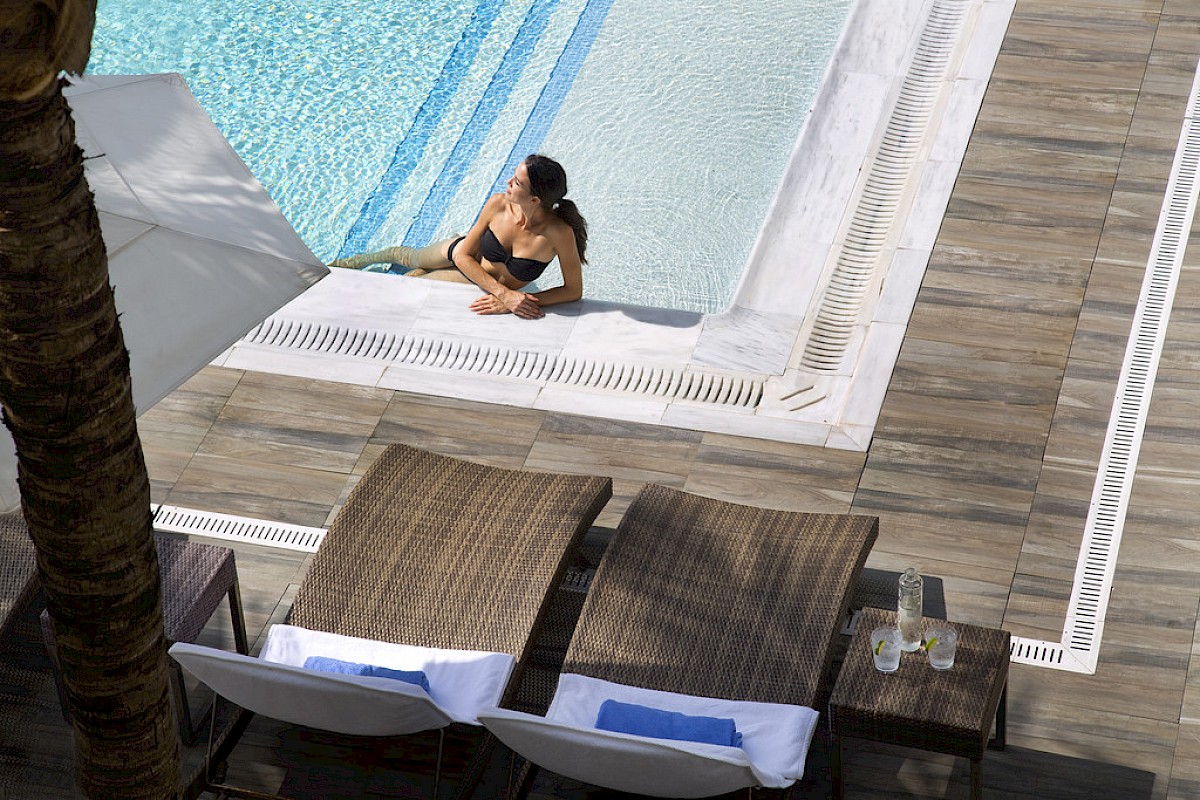 A person relaxes at the edge of a pool with two lounge chairs and towels nearby, under an umbrella.