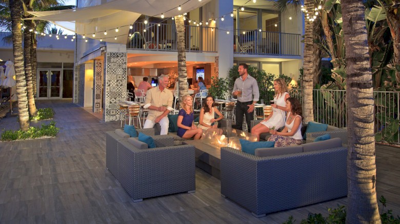 A group of people is relaxing on outdoor sofas around a fire pit, with string lights above and a building in the background.