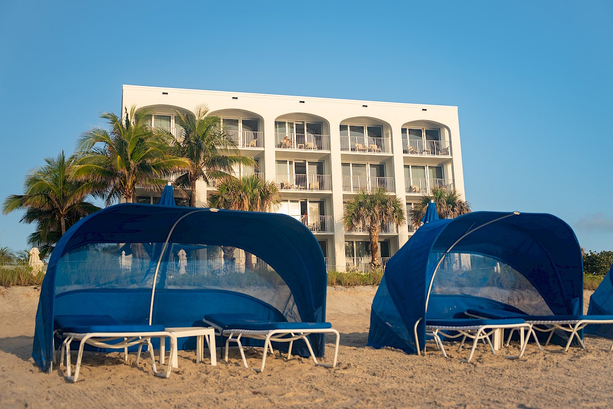 Beach scene with blue cabanas and lounge chairs on sand, in front of a white building with balconies and palm trees under a clear sky.