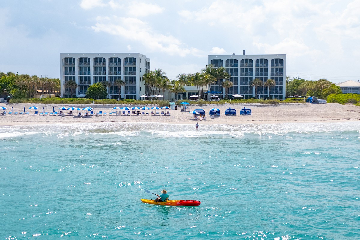 A person in a kayak is on the bright blue ocean near a sandy beach. Buildings and beach chairs line the shore under a cloudy sky.