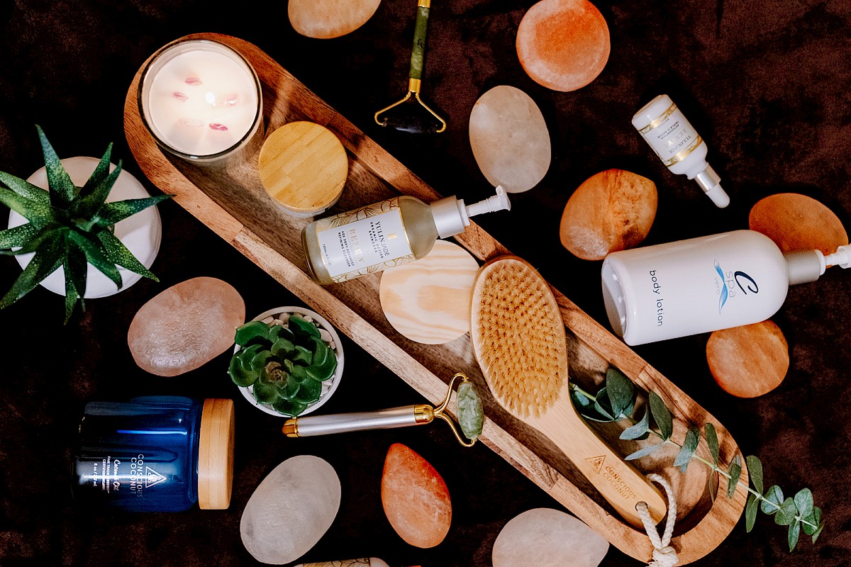 The image shows a flatlay of spa items like a candle, brush, jade roller, skincare bottles, and plants on a textured background.