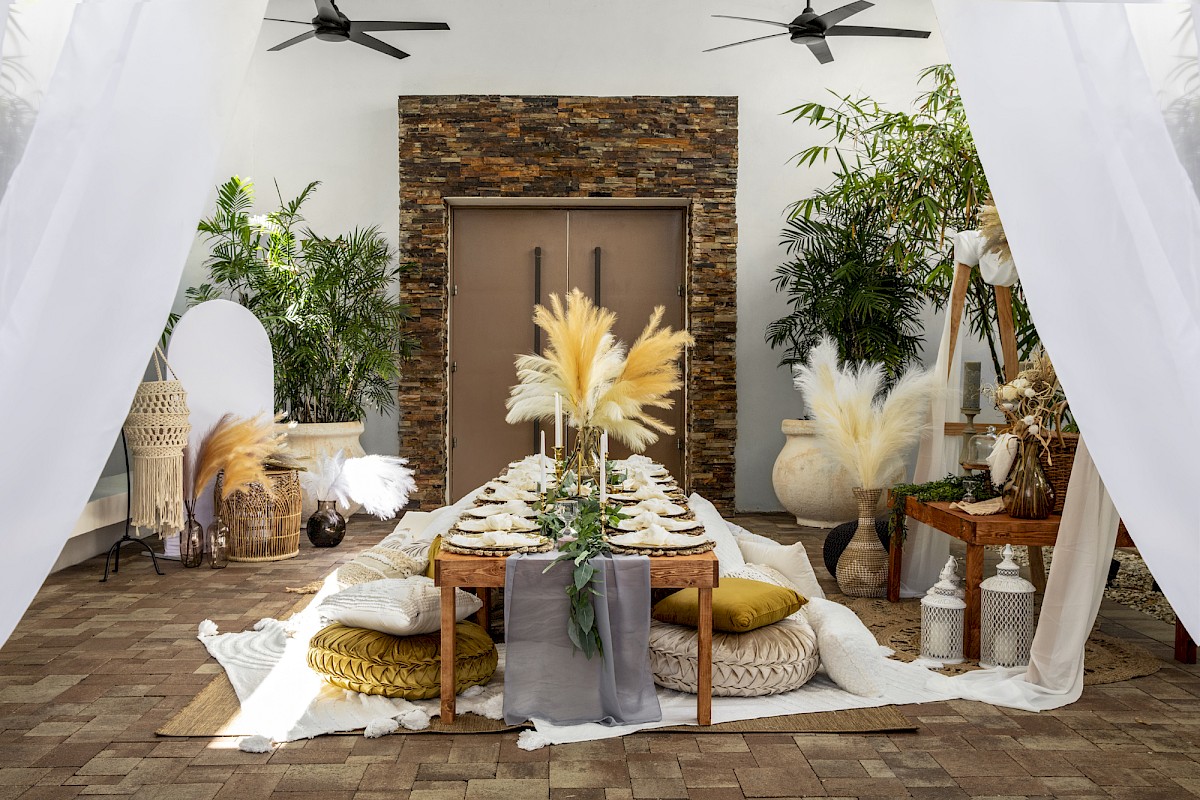 Elegantly set outdoor dining area with rustic decor, pampas grass centerpieces, pillows, and lush plants, under sheer white drapes.
