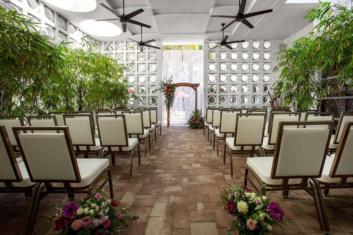 The image shows a wedding ceremony setup with chairs, floral arrangements, and green plants in a bright, open space.