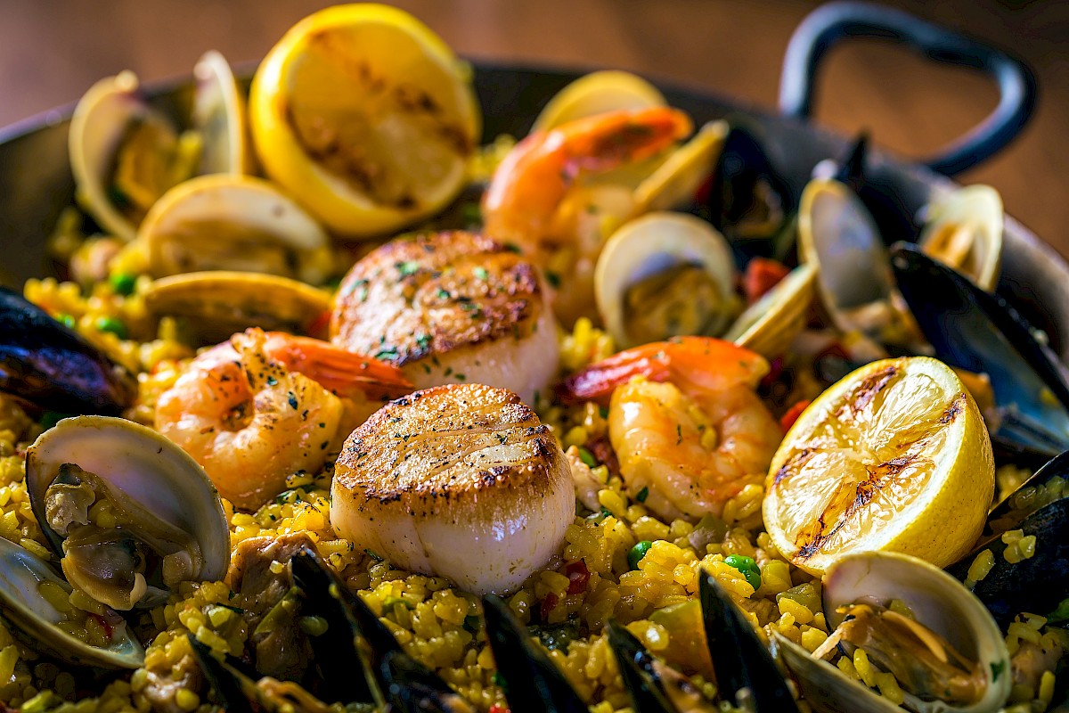 A seafood paella with rice, shrimp, scallops, clams, mussels, and lemon slices, all cooked in a large pan, garnished with herbs.