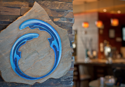 A stone plaque with a blue, circular wave design is on a wall, with a blurred interior and hanging lights in the background.