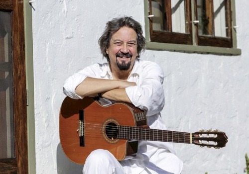 A person is smiling, sitting outside with an acoustic guitar, leaning against a white wall and near wooden-framed windows.