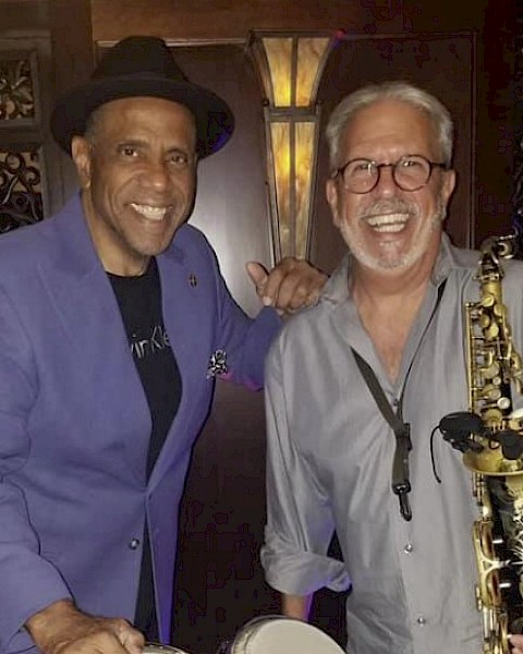 Two smiling men, one holding bongos, the other a saxophone, in a music-themed setting, wearing casual attire.