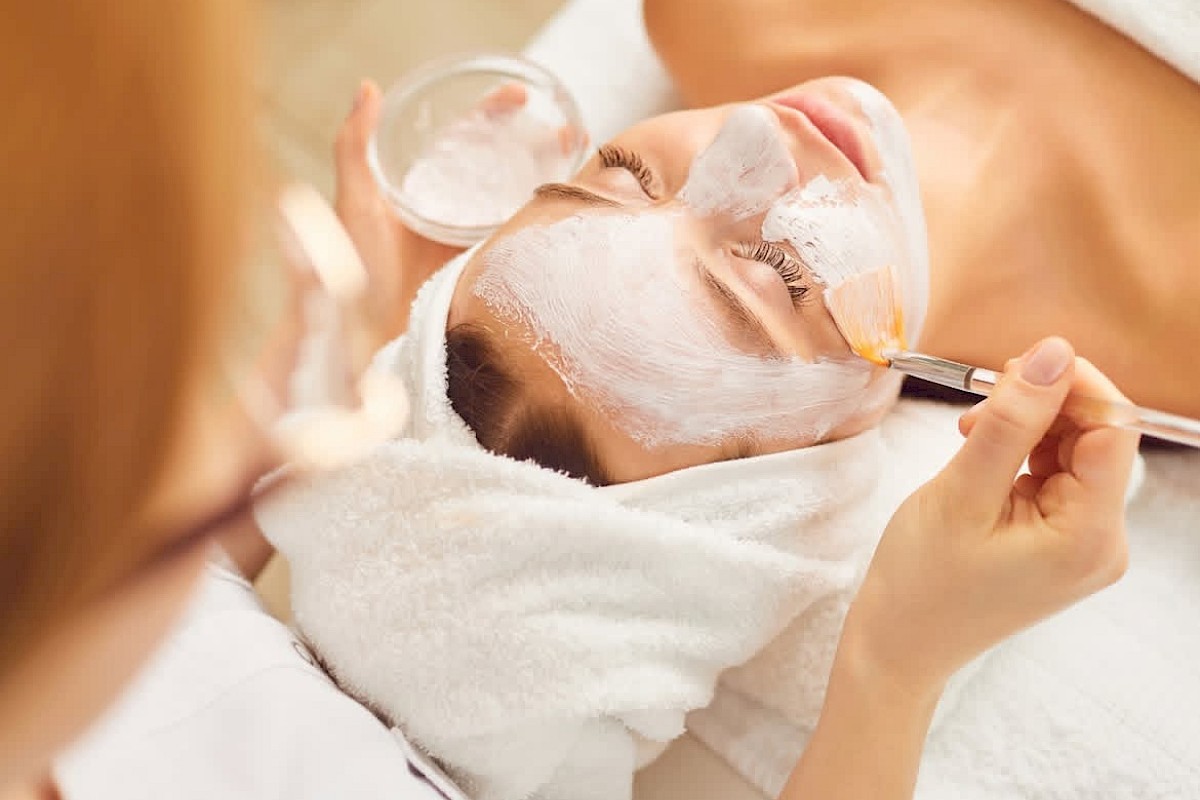 A person is receiving a facial treatment. Cream is being applied to their face with a brush as they lie down with a towel on their head.