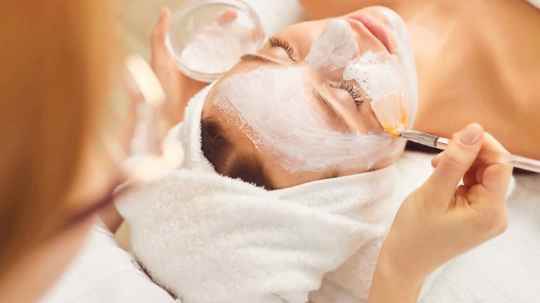 A person is receiving a facial treatment. Cream is being applied to their face with a brush as they lie down with a towel on their head.