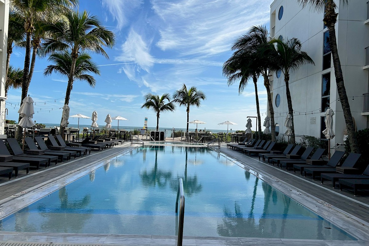 A serene outdoor pool surrounded by lounge chairs and palm trees, under a clear blue sky with wispy clouds near a white building.