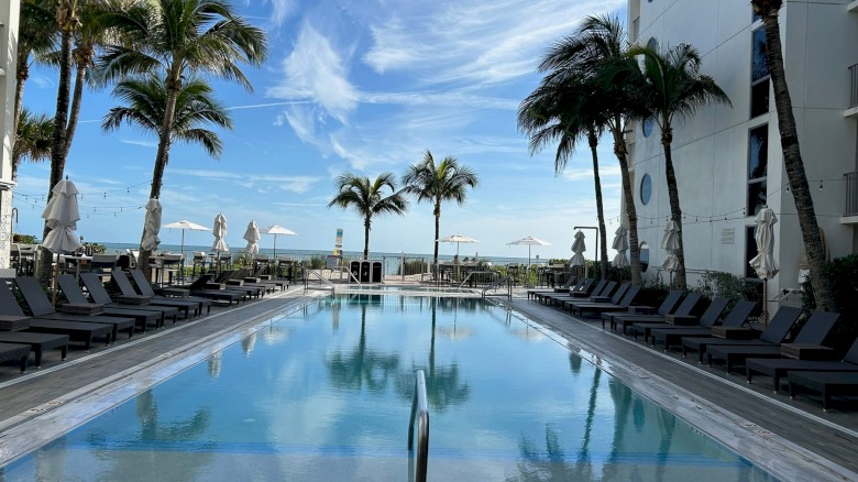 A serene outdoor pool surrounded by lounge chairs and palm trees, under a clear blue sky with wispy clouds near a white building.