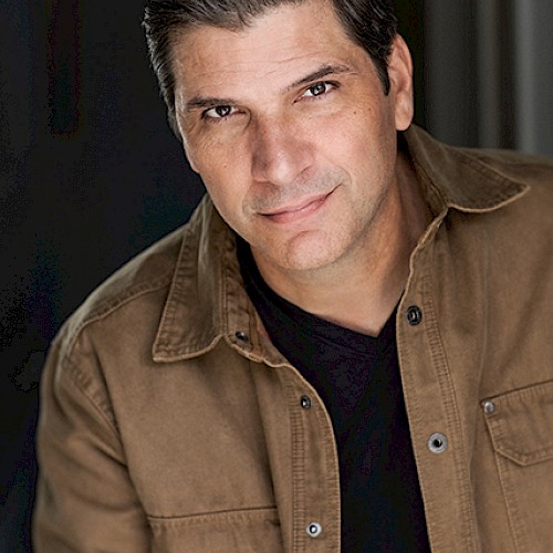 A person in a brown jacket over a black shirt is looking at the camera, set against a dark background.