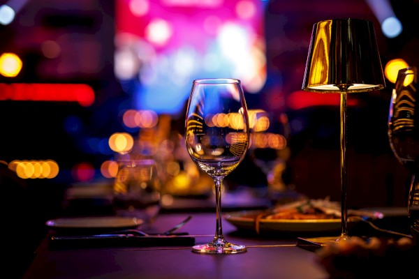 A dimly lit restaurant table with a wine glass, cutlery, and a small lamp, set against a blurred colorful background of lights.