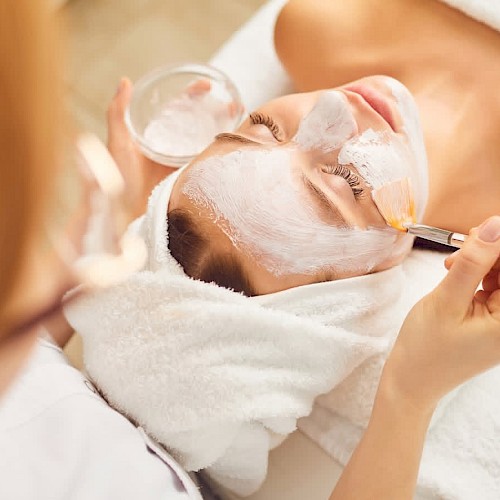 A person is receiving a facial treatment with a brush applying a mask, while lying down with a towel wrapped around their head.