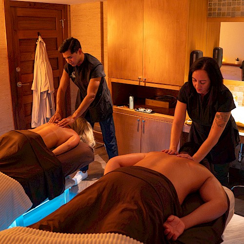 Two people receiving massages in a cozy room, with therapists providing treatment under warm lighting.