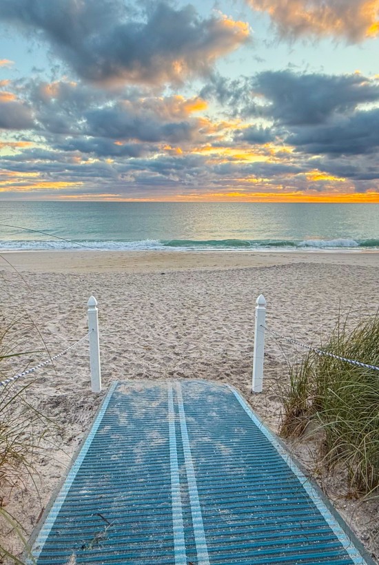 A boardwalk leads to a sandy beach with grass on the sides, under a cloudy sky and colorful sunset over calm ocean waves.